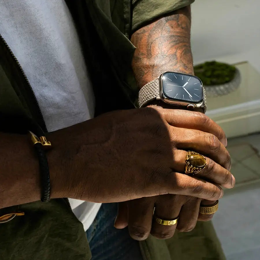 Nahaufnahme eines männlichen Handgelenks mit tätowiertem Arm, das das Arqura Apple Watch Armband in Clay trägt. Gewebtes Nylon mit D-Ring-Verschluss, kombiniert mit goldfarbenem Schmuck und modernem Streetstyle-Look im Sonnenlicht.