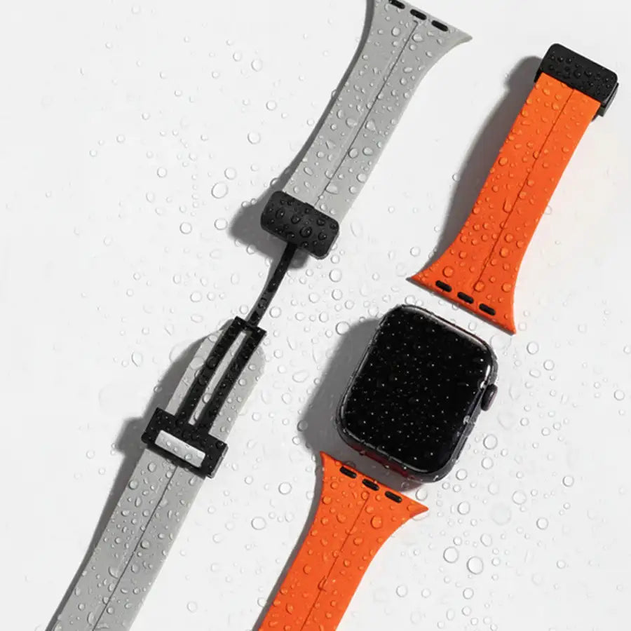 Zwei wasserfeste Linéa Apple Watch Armbänder in Grau und Orange mit schwarzem Magnetverschluss. Sportliches, modernes Design für aktive Träger:innen, abgebildet mit Apple Watch auf nasser Oberfläche.